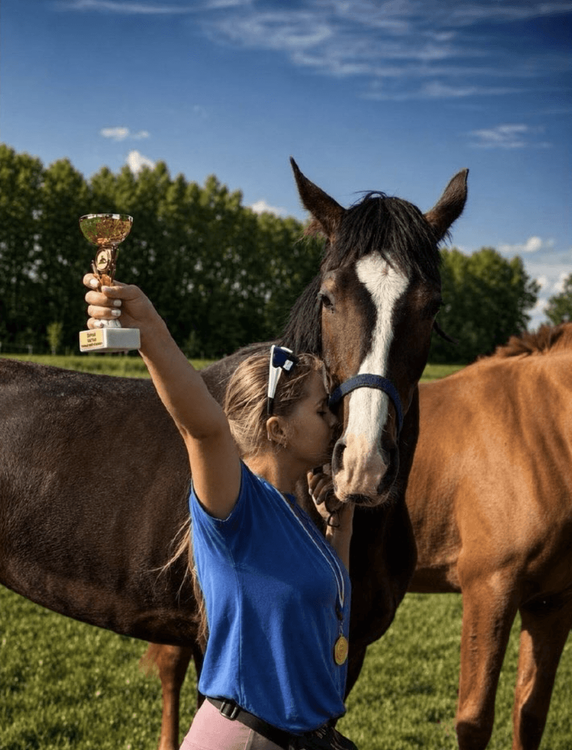 Equestrian rider proudly holds a gold trophy next to her horse, celebrating their victory.